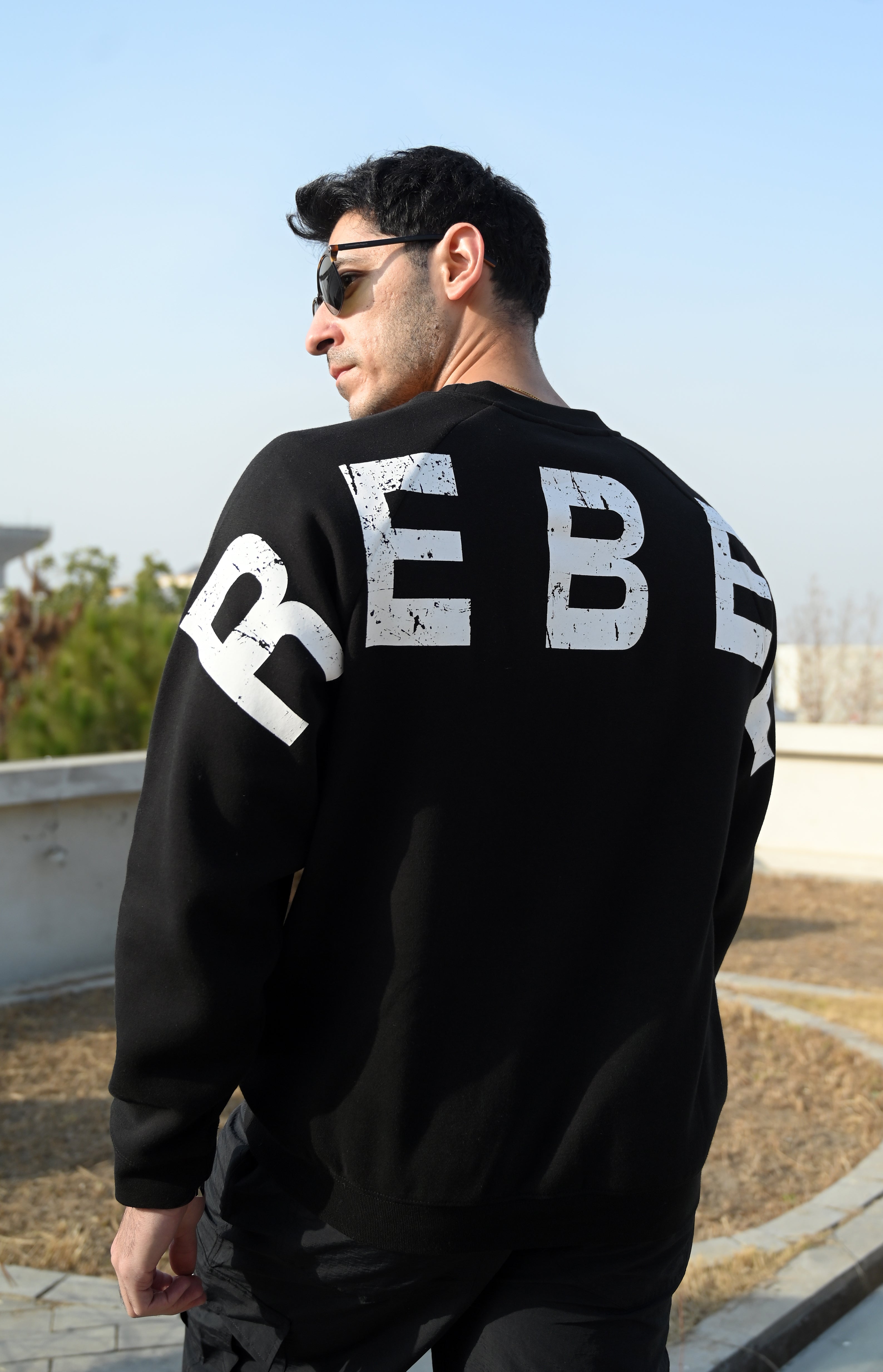 Person wearing a black sweatshirt with 'REBEL' printed on the back outdoors.