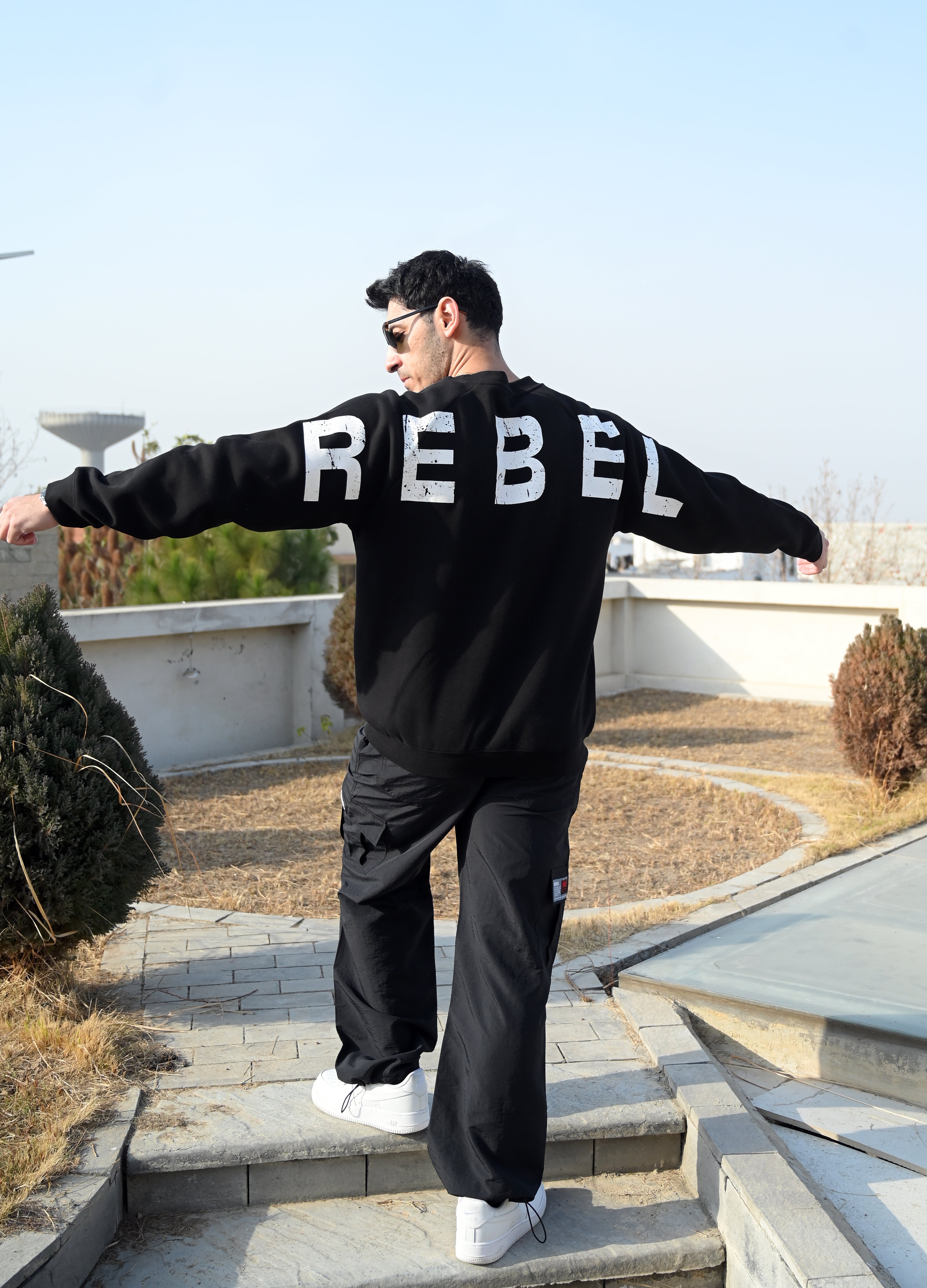 Person wearing a black hoodie with 'REBEL' printed on the back, standing outdoors.