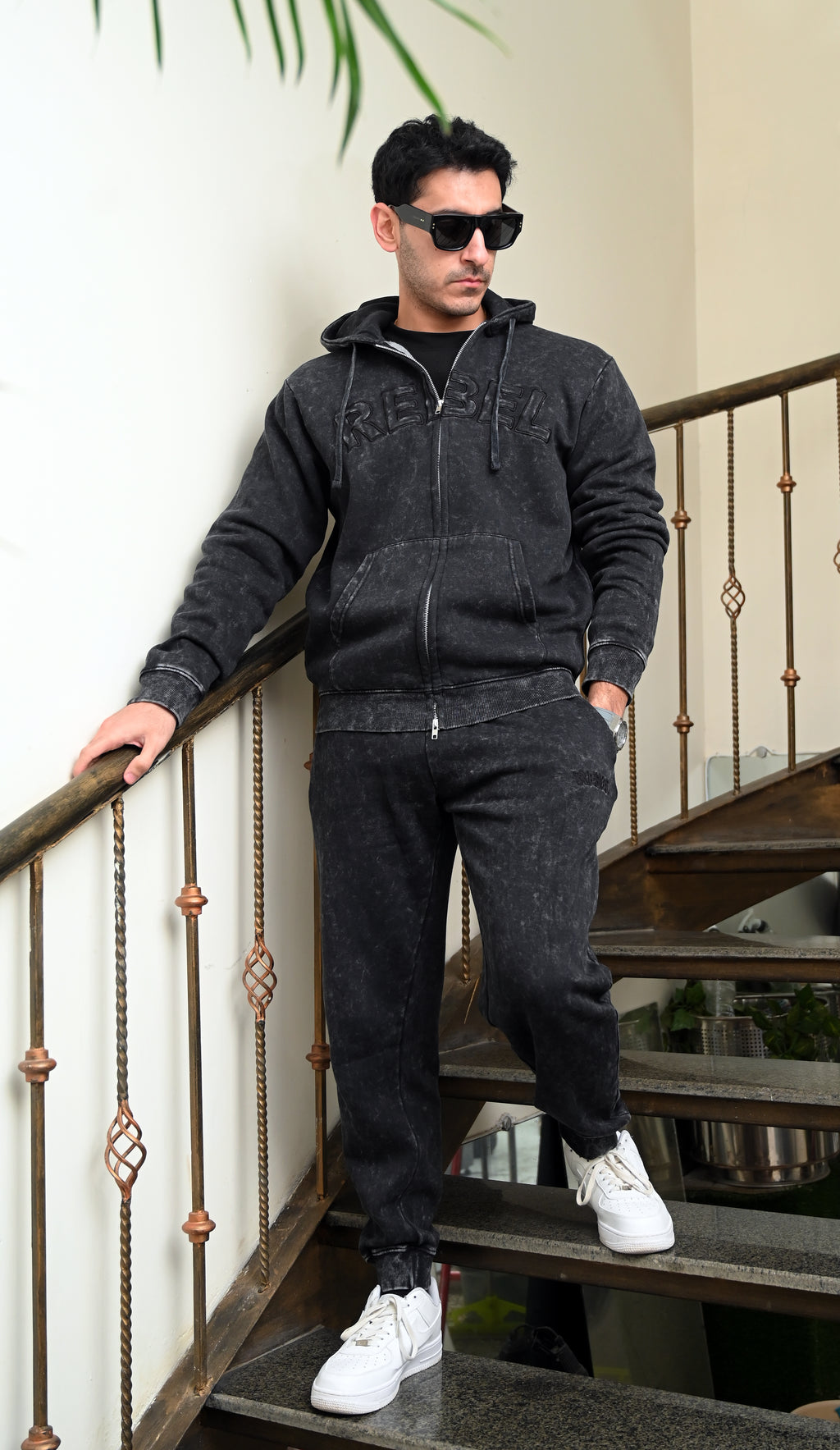 Man wearing a dark gray hoodie and pants with sunglasses, standing on a staircase.