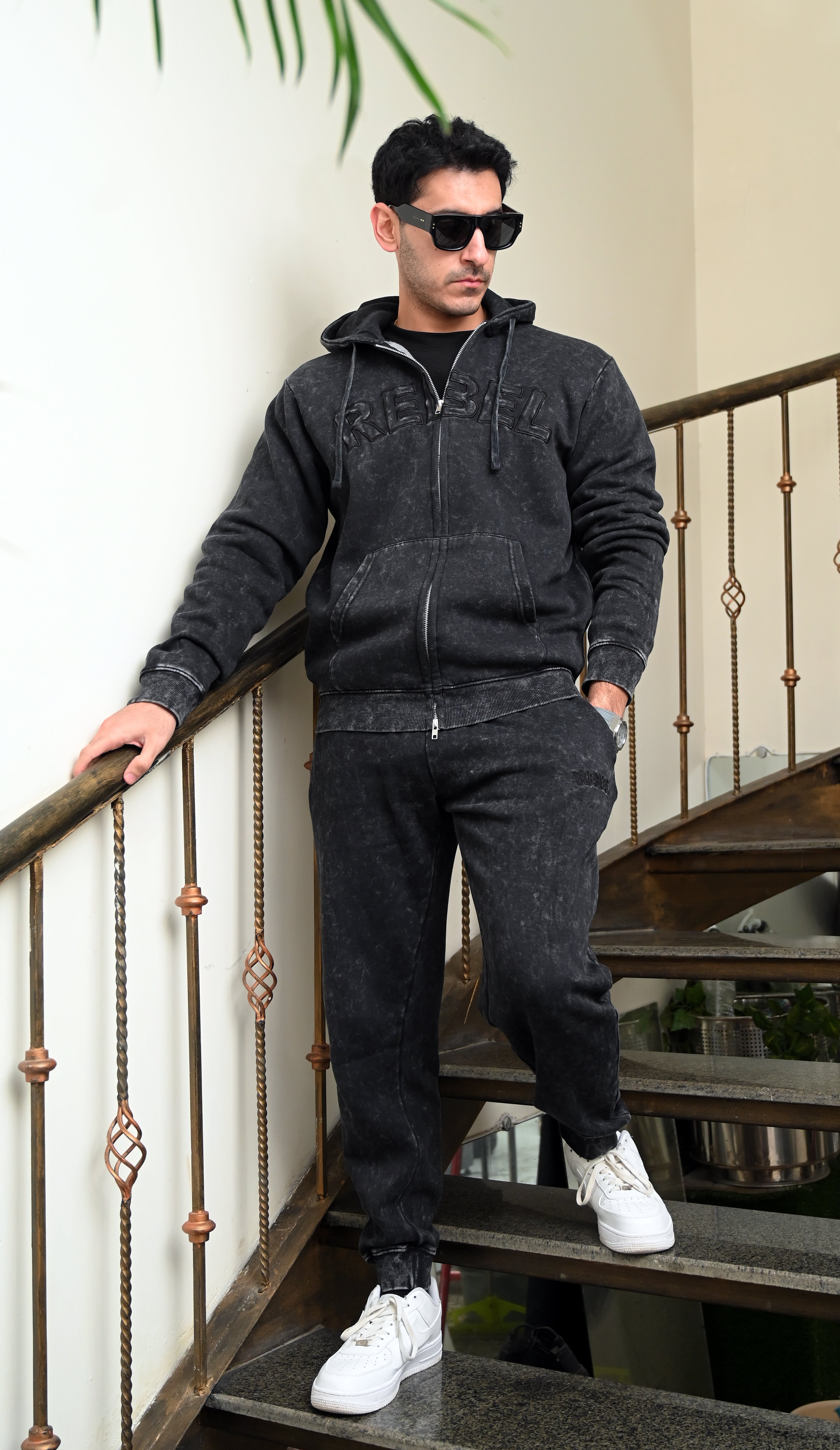 Man wearing a dark gray hoodie and pants with sunglasses, standing on a staircase.