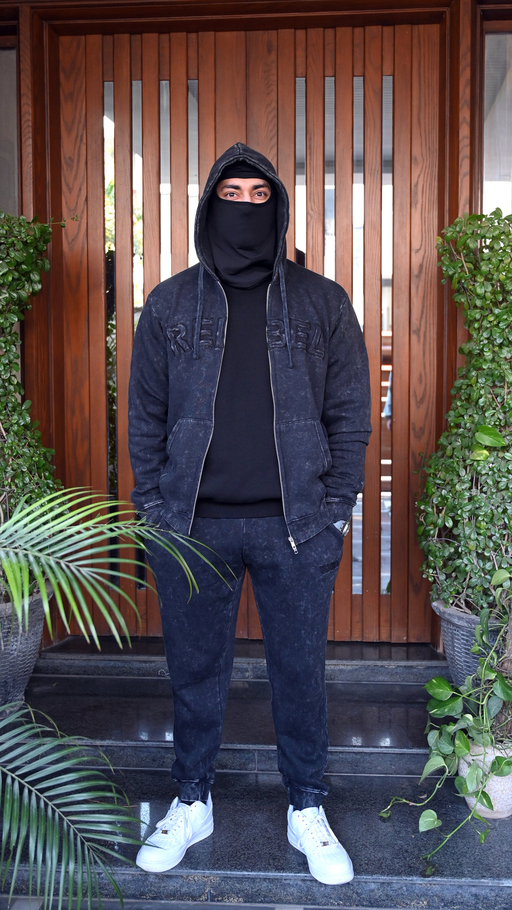 Person wearing a dark outfit with a hood and face covered, standing in front of a wooden door.