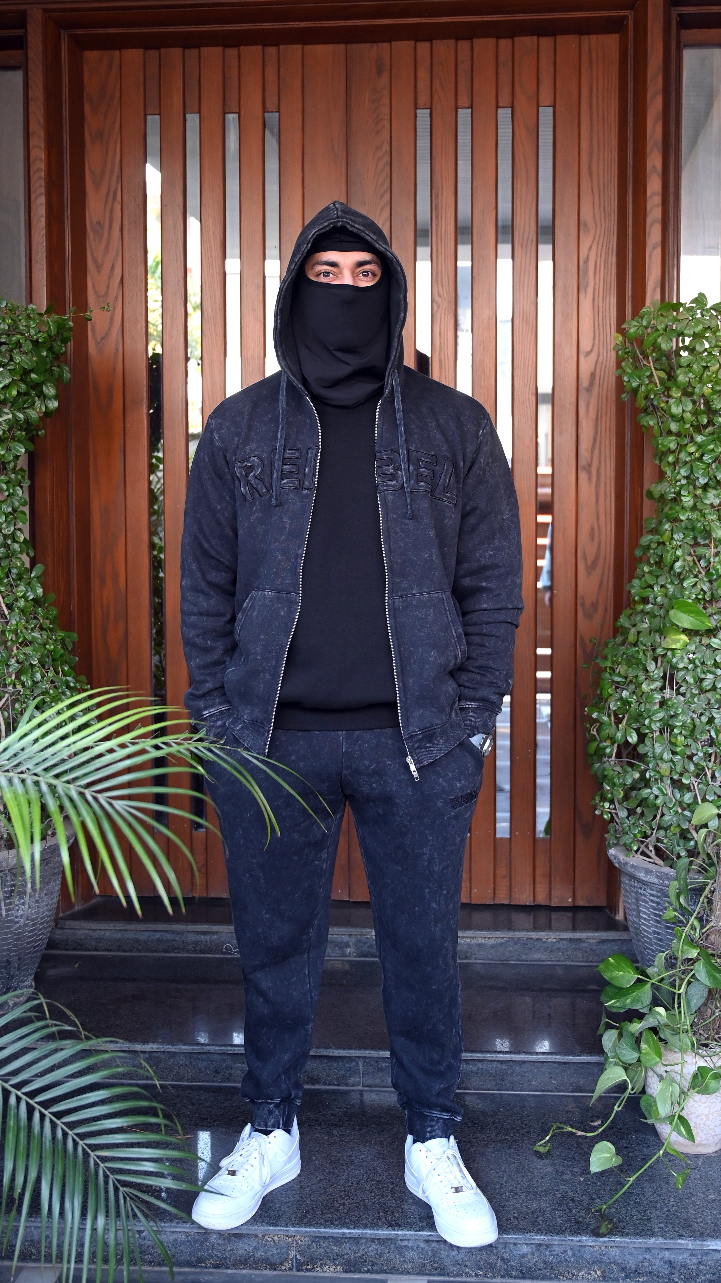 Person wearing a dark outfit with a hood and face covered, standing in front of a wooden door.