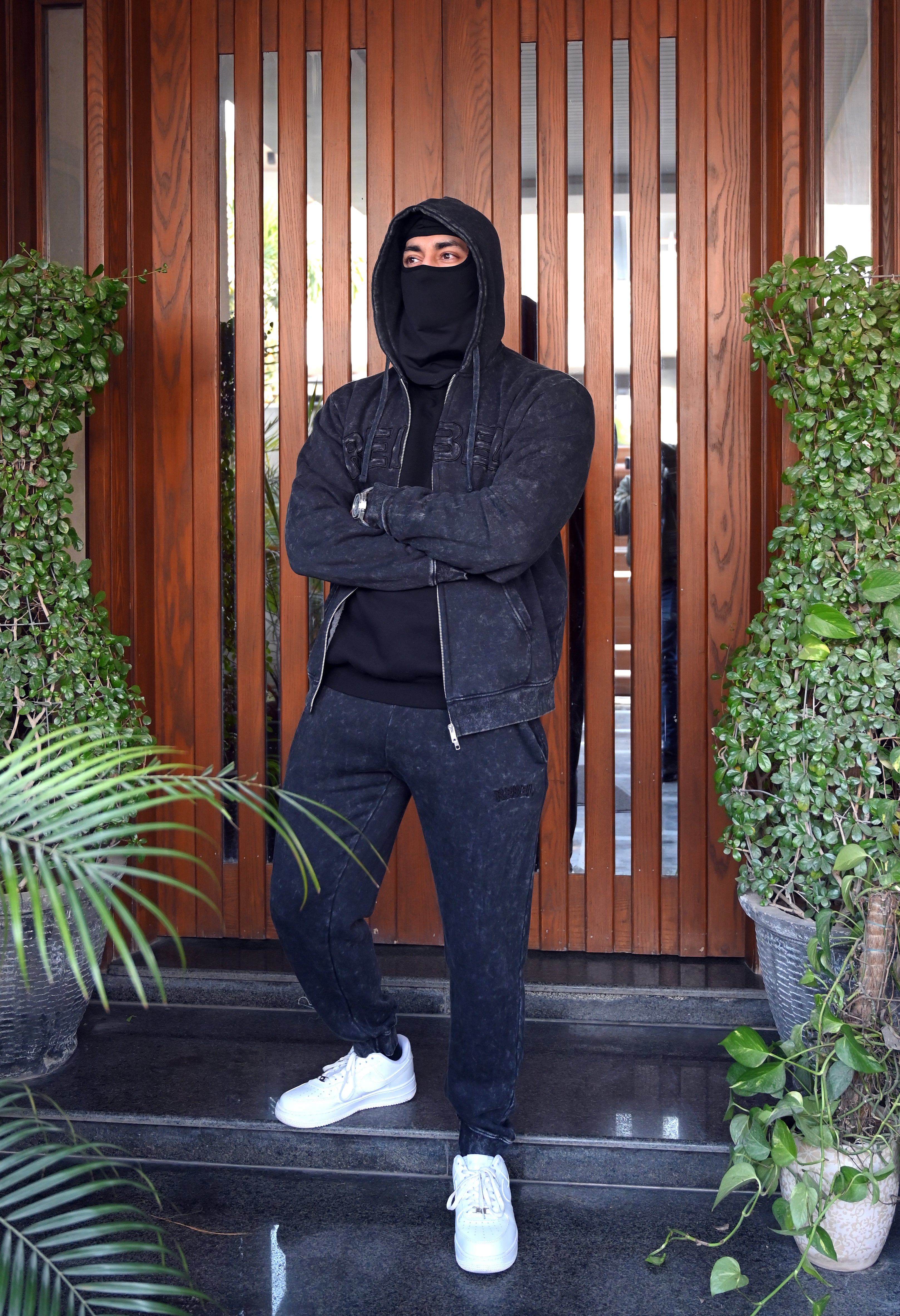 Person wearing a black hoodie and pants standing in front of a wooden gate with plants on either side.